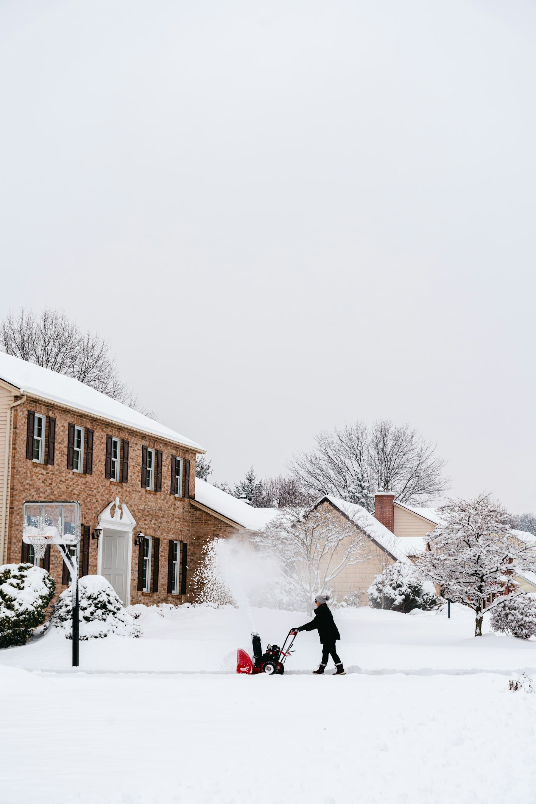 A person clearing a snowy driveway with a snow blower in front of a brick house