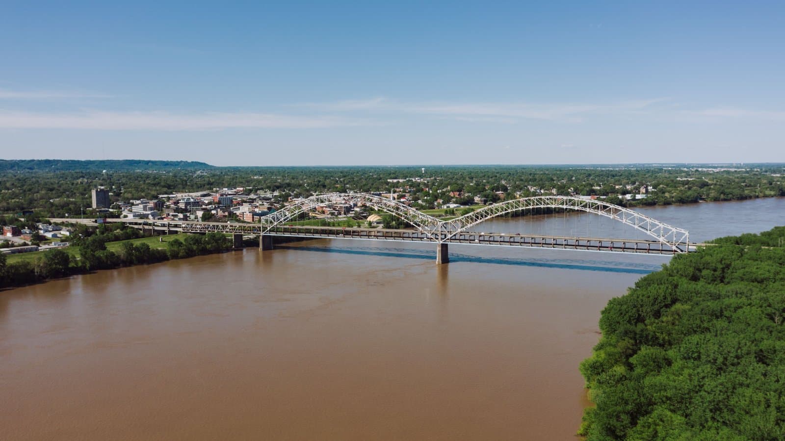 A scenic aerial view of a bridge crossing a river in Louisville, KY, with lush greenery and a clear blue sky.
